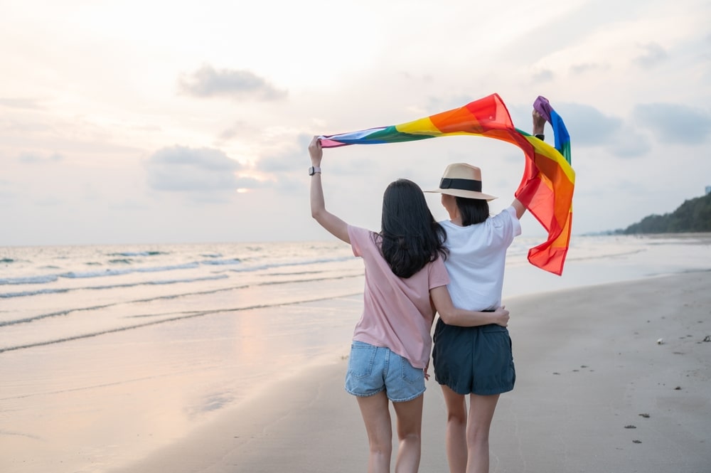 Zwei Frauen in der Rückansicht am Strand, sie halten eine Pride-Flagge hoch